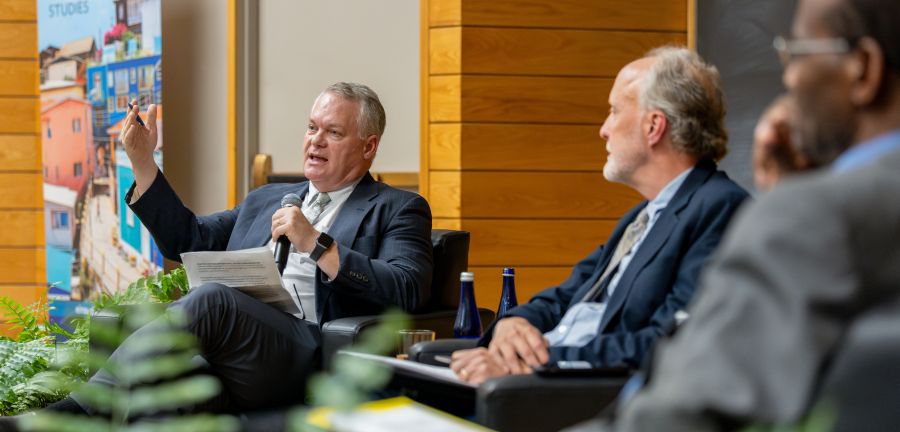Chris Barrett speaks into a microphone next to Paul Kaiser and Muna Ndulo.