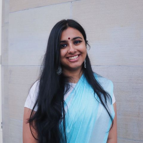 Pritika, wearing blue, stands in front of a grey wall. 