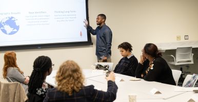 Obioha Chijioke speaks to a small group while pointing toward a presentation slide.